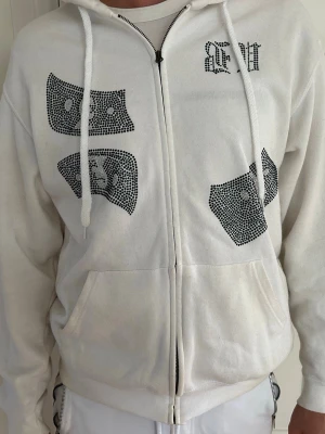 Vit hoodie med stenar och tryck  - Vit zip-hoodie med luva och dragsnören från Bvaldi, dekorerad med svarta strasstenar som formar sedlar och texten 'Make it rain' på ryggen. Köpt från Bvaldi på väla i Helsingborg.