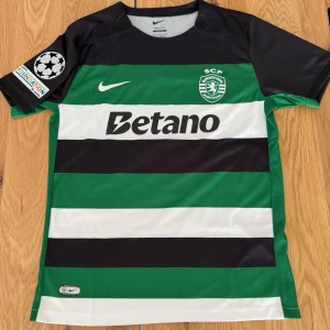 Sporting CP matchtröja Gyökeres #9 - Sporting CPs officiella matchtröja från Nike med gröna och vita horisontella ränder, svarta detaljer och sponsortryck. Nummer 9 och namnet Gyökeres på ryggen. Champions League-märke på ärmen. Perfekt för fotbollsfans!