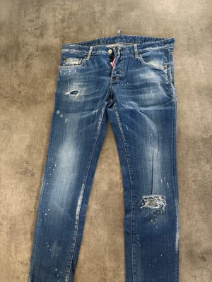 Blå jeans från Dsquared2 - Säljer ett par blå jeans från Dsquared2 med distressed detaljer, slitningar och färgstänk. Jeansen har smal passform, klassisk femficksmodell och knappgylf. Perfekta för en edgy och trendig look.