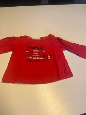 Röd långärmad blus med tryck - Söt röd tröja med lång ärm och rund halsringning. Framsidan har ett lekfullt tryck med text och små motiv. Tröja har en avslappnad passform och är perfekt för en färgglad vardagslook.