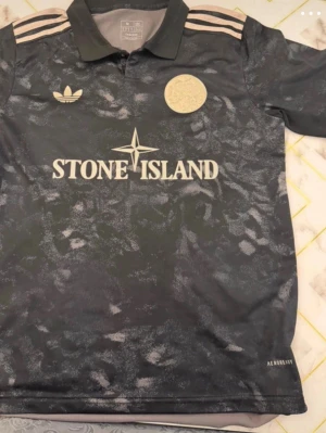 Svart Ajax matchtröja Stone Island x Adidas - Svart sporttröja med grått mönster, klassisk krage och kort ärm. Framtill syns Stone Island-logga och Adidas-märke i vitt. Tröjan har tre ränder på axlarna och är tillverkad i lätt, ventilerande material med Aeroready-teknologi.