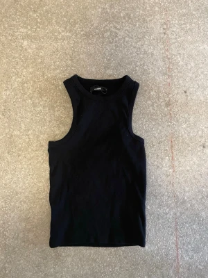 Svart ribbad linnetopp från bikbok  - En svart, ärmlös linnetopp från bikbok i ribbad bomull. Toppen har rund halsringning och en tight passform som sitter snyggt mot kroppen. Perfekt att styla med jeans eller kjol för en clean look.
