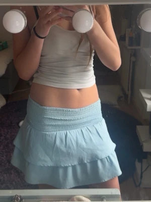 Ljusblå volangkjol med smock - Supersöt ljusblå kortkjol med bred smock i midjan och dubbla volanger. Kjolen har en flowy känsla och passar perfekt till sommaren. Enkel att matcha med crop tops eller linnen för en chill look.
