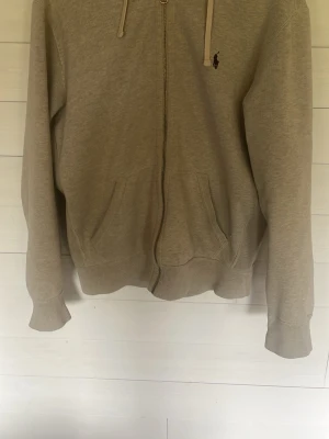 Beige hoodie från Polo Ralph Lauren - Säljer en beige hoodie från Polo Ralph Lauren med dragkedja framtill och två fickor. Klassisk huva med snören och den ikoniska lilla loggan broderad på bröstet. Tillverkad i mjukt bomullsmaterial, perfekt för en avslappnad och stilren look.