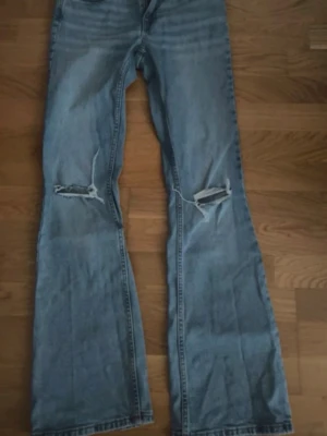 Ljusblå bootcut jeans med slitningar - Ljusblå bootcut jeans från 157 med slitna detaljer och hål vid knäna. Klassisk femficksmodell med dragkedja och knapp. Perfekta för en avslappnad och trendig look.