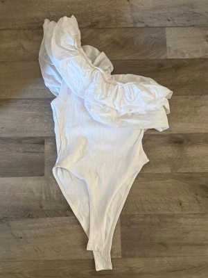 Volangbody Zara - Snygg vit baddräkt med asymmetrisk design och stor volang över ena axeln. Tillverkad i ribbat material som ger en modern känsla och sitter skönt på kroppen. Perfekt för dig som vill ha en unik och trendig look på stranden.