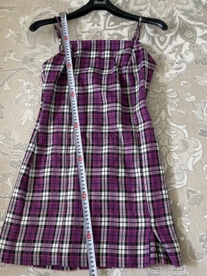 Lila rutig kortklänning med slits - Snygg kortklänning med rutigt mönster i lila, svart och vitt. Klänningen har smala axelband, rak halsringning och en liten slits framtill. Och dragkedjan bak på ryggen. Perfekt för dig som gillar färg och mönster. Stängs med dragkedja i ryggen.