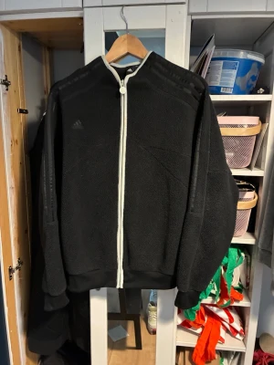 Svart fleecejacka från Adidas - Mysig svart fleecejacka från Adidas med hel dragkedja framtill i vitt och diskret Adidas-logga på bröstet. Jackan har långa ärmar, hög krage och ribbade muddar. Perfekt för lager på lager och chill dagar.