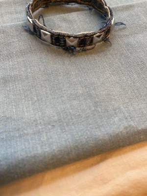 Jeansarmband med nitar - Snyggt armband i denimtyg med silvriga fyrkantiga nitar. Armbandet har en cool, ruffig look med fransiga kanter och passar perfekt för dig som gillar en edgy stil. Justerbar storlek med knytband.