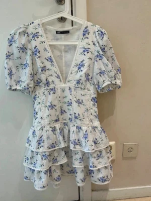  Ny trendig Vit blommig volangklänning från Zara - Superfin kort klänning från Zara i vitt med blått blommönster. Klänningen har djup v-ringning, puffärmar och flera volanger nertill. Tillverkad i bomull och perfekt för sommarens alla tillfällen. Söt spetskant längs urringningen ger extra detalj.