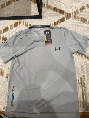 Grå t-shirt från Under Armour - Säljer en grå t-shirt från Under Armour med korta ärmar och tryckt logga på bröstet. Materialet är lätt och andas, perfekt för träning. Färgade detaljer och texten UNDER ARMOUR längs sidan ger en cool look. Passar dig som vill ha något stilrent och sportigt.