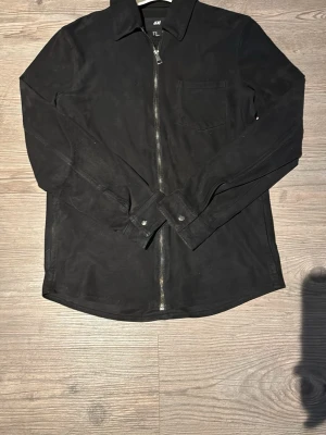 Svart overshirt med dragkedja H&M - Stilren svart overshirt från H&M med hel dragkedja framtill, klassisk krage och en bröstficka. Långärmad modell med knappar vid ärmslut. Perfekt att slänga över en t-shirt för en clean look.
