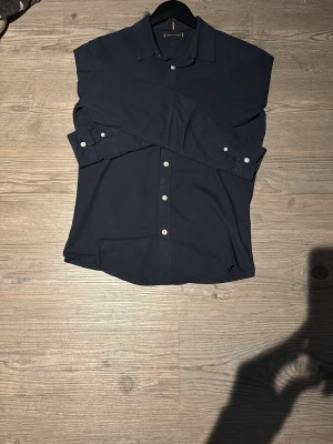 Mörkblå skjorta från Tommy Hilfiger - Klassisk mörkblå skjorta från Tommy Hilfiger med vita knappar och diskret logga på bröstet. Skjortan har lång ärm, krage och är tillverkad i bomull. Perfekt för en stilren look.