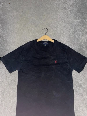 Svart t-shirt från Polo Ralph Lauren - Svart t-shirt från Polo Ralph Lauren med klassisk passform och korta ärmar. Liten röd broderad logga på bröstet. Perfekt till jeans eller shorts för en stilren look.