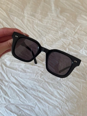 Chimi glasögon  - Klassiska svarta solglasögon från Ray-Ban med rund form och tunna metallbågar. Glasen är mörka och ger en stilren vibe, perfekt för soliga dagar. En ikonisk accessoar som alltid är trendig.