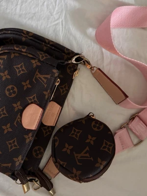 axelväska med monogram - Säljer en brun axelväska  med klassiskt monogrammönster i beige och guldiga detaljer. Väskan har en bred rosa axelrem och en matchande rund coin purse som hänger på remmen. Tillverkad i canvas med läderdetaljer. Köpt i Turkiet. 