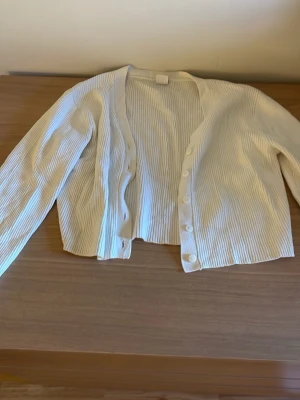 Vit ribbad kofta med knappar - Säljer en stilren vit kofta med ribbad struktur och långa ärmar. Koftan har en rak passform och knäpps framtill med vita knappar. Perfekt att slänga över en topp för en enkel och clean look.