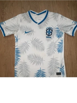 Vit Brasilien fotbollströja Nike - Snygg vit fotbollströja från Nike med Brasilien-emblem på bröstet. Tröjan har blå detaljer vid krage och ärmslut samt ett mönster av palmblad i blått och grått. Perfekt för dig som gillar sportig stil och vill visa ditt stöd för Brasilien.