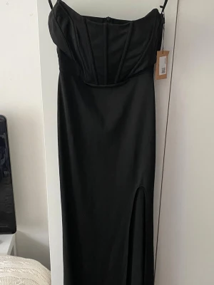Svart lång klänning med slits - Snygg svart lång klänning och markerad byst. Klänningen har en figurnära passform och en hög slits framtill. Materialet är glansigt och ger en elegant look. Köpt som balklänning från sellpy men då jag hitta en annan säljer jag nu denna☺️