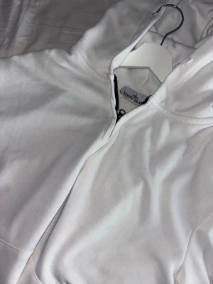 Vit hoodie från Stone Island - Snygg vit hoodie från Stone Island med dragkedja och huva. Tillverkad i mjukt bomullsmaterial som känns skönt mot huden. Perfekt för en clean och stilren look. Klassisk passform och diskret logga i nacken.
