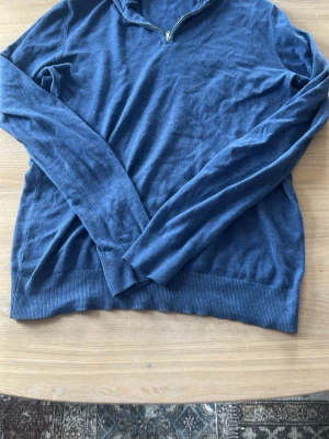 Blå tunnstickad tröja med halvzip - Mysig blå tunnstickad tröja med halv dragkedja vid halsen. Långärmad modell med ribbade muddar och nederkant. Perfekt för lager med 100% bomull.