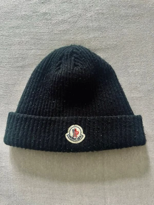 Svart ribbstickad mössa  - Svart ribbstickad mössa med klassisk uppvikt kant och broderad logotyp framtill. Tillverkad i mjukt material som håller dig varm och ger en stilren look. Perfekt accessoar för kyliga dagar.