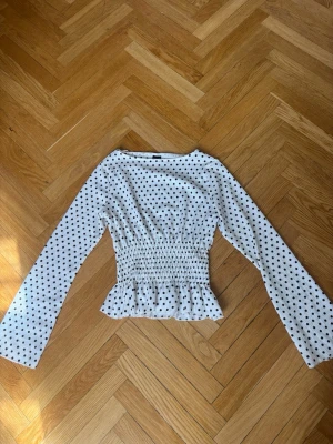 Vit prickig blus från Gina Tricot - Supersöt vit blus med svarta prickar från Gina Tricot. Modellen har långa ärmar, smockad midja och volangkant nedtill. Säljer då den inte sitter så bra på mig och därför inte kommer till användning. Jag har använt den typ 2 gånger max🌸