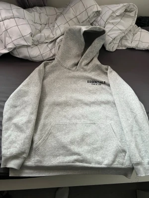 Grå Essentials Fear of God hoodie - Snygg grå hoodie från Essentials Fear of God med svart logga på bröstet. Klassisk modell med stor huva och magficka. Perfekt för dig som gillar streetwear och vill ha en stilren look. Mjuk och skön bomullsblandning.