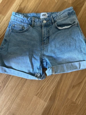 Blå denimshorts från lager 157 - Säljer ett par klassiska blå denimshorts från lager 157 med upprullade benslut och femficksmodell. Shortsen har normal passform och är tillverkade i bomull. Perfekta för sommaren och enkla att matcha med olika stilar.