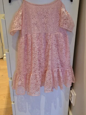 Ljusrosa spetsklänning med volang - Söt ljusrosa kortklänning i spets med volangdetaljer och korta ärmar. Klänningen har ett romantiskt uttryck med genombrutet mönster och en luftig passform. Perfekt för sommarens alla tillfällen.