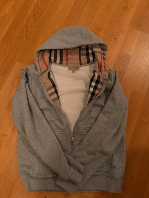 Grå hoodie från Burberry med dragkedja - Snygg grå hoodie från Burberry med klassiskt rutigt foder i huvan och dragkedja framtill. Mjuk insida i fleece och två fickor. Perfekt för dig som gillar stilrena plagg med exklusiv känsla och ikoniska detaljer.