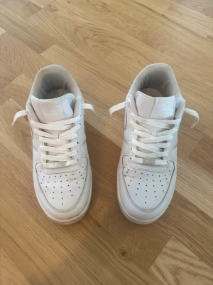 Nike Air Force 1 vita sneakers - Klassiska Nike Air Force 1 sneakers i helvitt läder med perforerad tåbox och vita skosnören. Snygg och tidlös modell med metallbricka märkt 'AF1' på snörningen. Perfekta för en clean och stilren look.
