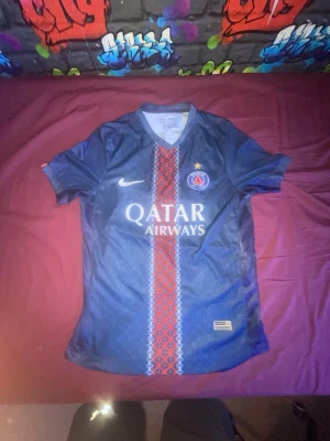 PSG matchtröja Nike blå/röd - Säljer en blå Paris Saint-Germain matchtröja från Nike med röd och vit rand längs mitten, PSG-logga och Qatar Airways-tryck på bröstet. Tröjan har korta ärmar och sportig passform, perfekt för fotboll eller träning.