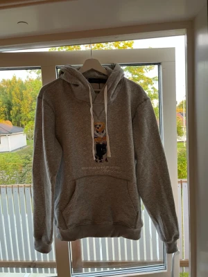 Grå hoodie Polo Ralph Lauren björn - Snygg grå hoodie från Polo Ralph Lauren med klassisk björn-tryck på bröstet. Perfekt för en avslappnad och trendig stil. Mjuk bomullsblandning och ribbade muddar. 