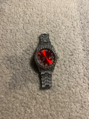 Silverfärgad klocka med röd urtavla - Snygg klocka med glittrande silverfärgat armband och boett, täckt av små stenar. Urtavlan är metallicröd och har tydliga markeringar samt datumfönster. Perfekt accessoar för dig som vill sticka ut med stil.