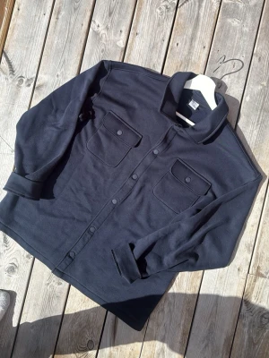 Svart overshirt med tryckknappar - Stilren svart overshirt med två bröstfickor med lock och tryckknappar. Skjortkrage och raka ärmar som kan vikas upp för en avslappnad look. Tillverkad i ett mjukt, tjockare tyg som ger en schysst lager-på-lager känsla.