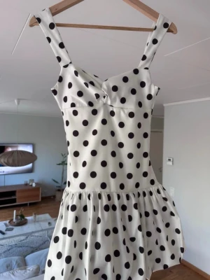  Ny trendig Vit klänning med svarta prickar - Supersöt vit kortklänning med svarta prickar och breda axelband. Klänningen har en lätt utställd kjol och markerad midja, perfekt för en trendig och lekfull look. Materialet känns mjukt och svalt, perfekt för varmare dagar.