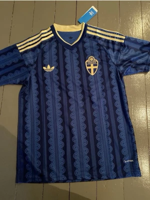 Blå Sverige fotbollströja Adidas - Retroinspirerad blå fotbollströja från Adidas med Sveriges landslagsemblem på bröstet. Tröjan har mönstrade vertikala ränder, gula detaljer vid krage och axlar samt Adidas-logga. Perfekt för dig som vill sticka ut på läktaren eller träningen.