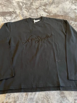 Svart sweatshirt från Acne Studios - Svart långärmad sweatshirt från Acne Studios med broderad logga framtill i matchande svart ton. Klassisk rund halsringning och avslappnad passform. Perfekt för en clean och stilren look.