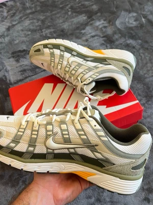 Nike P-6000 Light Army/Cargo Khaki/Light Orewood Brown/Phantom - Nike P-6000 sneakers med mesh och mockadetaljer i vitt, ljusgrått och olivgrönt. Swoosh-logga på sidan, gula detaljer vid sulan och en chunky retroinspirerad design. Perfekta för dig som gillar sportig och trendig stil.
