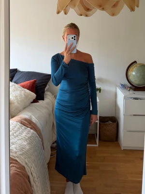 Blå oneshoulder långklänning - Snygg blå långklänning med oneshoulder-design och lång ärm. Klänningen har en smickrande drapering i midjan och är tillverkad i ett mjukt, stretchigt och lyxigt material som ger en bekväm passform. Perfekt för dig som vill sticka ut med en unik och elegant look.