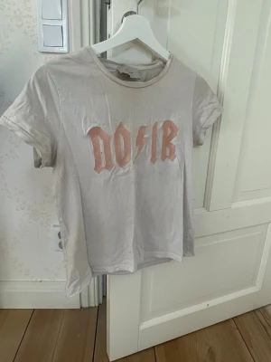 Vit t-shirt från Neo Noir med tryck - Vit t-shirt från Neonoir med coolt rosa tryck på bröstet där det står 'NOIR' med en blixt. Klassisk rund hals och korta ärmar. Perfekt till jeans eller kjol för en avslappnad look.