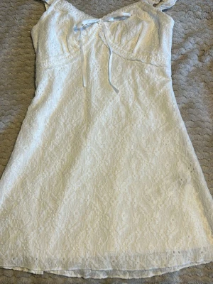 Vit spetsklänning med rosett - Romantisk vit kortklänning i spets med broderade detaljer och söt rosett framtill. Klänningen är ärmlös med volangkantade axelband och har en feminin, somrig känsla. Perfekt för dig som gillar ljusa och luftiga plagg. Säljer då jag kunde tyvärr ej returnera 💕