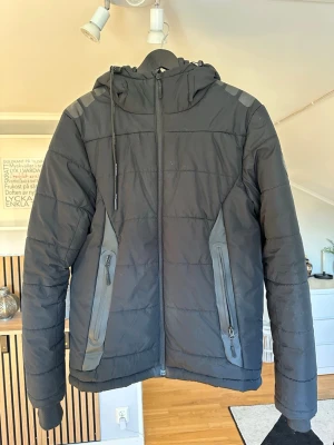 Svart pufferjacka från Bluewear - Snygg svart pufferjacka från Bluewear med huva och dragkedja framtill. Jackan har två stora snedställda fickor med dragkedja och detaljer i grått. Perfekt för kalla vinterdagar och har en sportig look.
