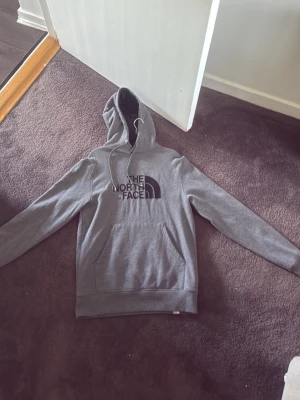 Grå hoodie från The North Face - Snygg grå hoodie från The North Face med stor svart logga på bröstet. Klassisk modell med huva och dragsko samt en stor magficka framtill. Perfekt för en avslappnad och sportig stil.