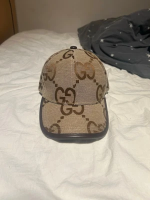 Beige keps med GG-mönster från Gucci - Säljer en beige keps från Gucci med det klassiska GG-monogrammet i brunt över hela kepsen. Kepsen har böjd skärm med mörkbrun kant och är tillverkad i canvasmaterial. Perfekt för dig som vill ha en stilren och lyxig accessoar.