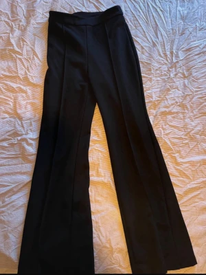 Svarta utsvängda kostymbyxor - Säljer ett par svarta kostymbyxor med hög midja och utsvängda ben. Byxorna har pressveck framtill för en clean look och är sydda i ett slätt tyg som ger en dressad känsla. Perfekta för dig som gillar stilrena och trendiga plagg.