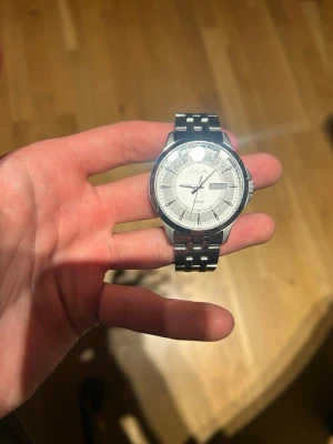 Citizen silverfärgad herrklocka - Stilren klocka från Citizen med rund silverfärgad boett och matchande länkarmband i metall. Urtavlan är ljus med tydliga silverfärgade markeringar och visare samt dag- och datumvisning. Perfekt accessoar för dig som gillar klassisk och clean stil.