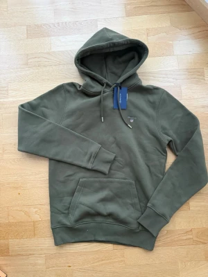 Olivgrön hoodie från GANT med prislapp kvar - Snygg olivgrön hoodie från GANT med klassisk huva och dragsko. Framtill finns en känguruficka och GANT:s logga broderad på bröstet. Tillverkad i mjuk bomullsblandning som känns skön mot huden. Perfekt för en avslappnad och trendig look. Prislappen kvar då den aldrig blivit använd och nu är den för liten för min son. 