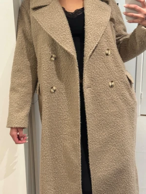 Beige teddy-kappa med dubbelknäppning - Snygg beige teddy-kappa med dubbelknäppning och stora slag. Kappan har två fickor med lock och en rak, lång passform. Tillverkad i mjukt, fluffigt material som ger en trendig look. Perfekt för dig som vill ha en varm och stilren ytterjacka. Oversized jacka passar M och L beror på hur man vill att den ska sitta.